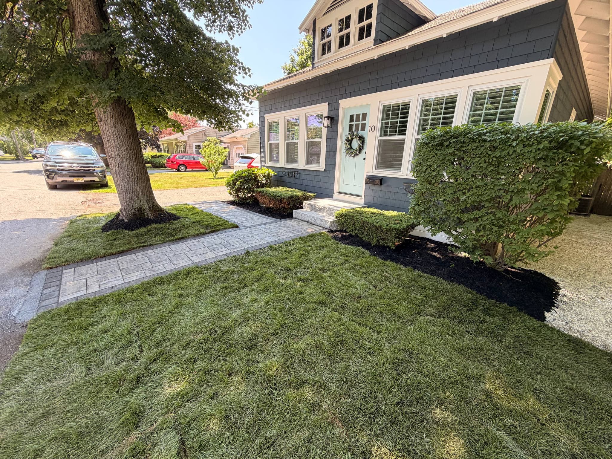 Angled view of the completed entry showing the paver walkway, granite steps, and newly sodded front lawn beneath a shade tree