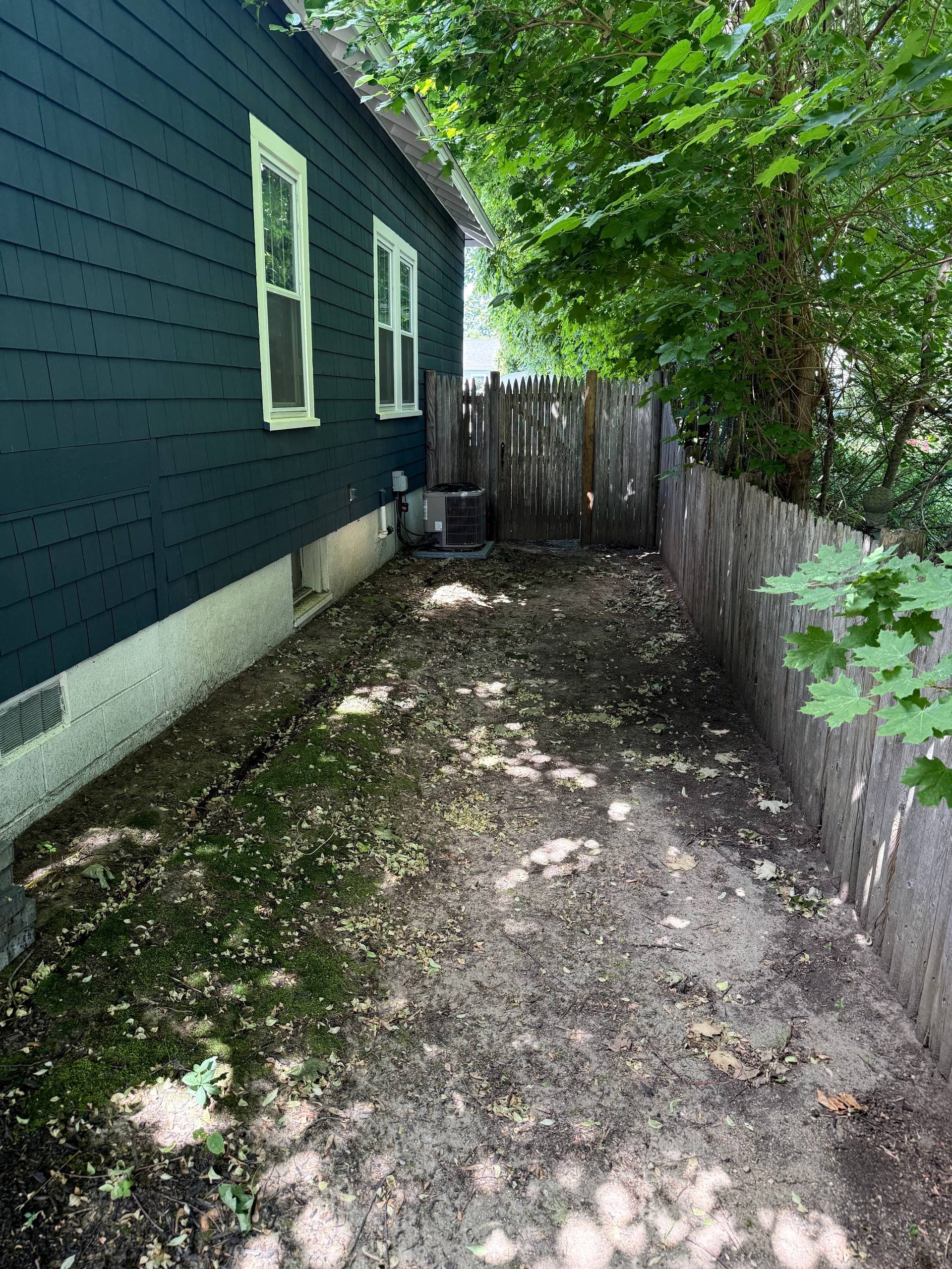 Original side yard before the rebuild — bare dirt and leaf-covered ground running along the fence
