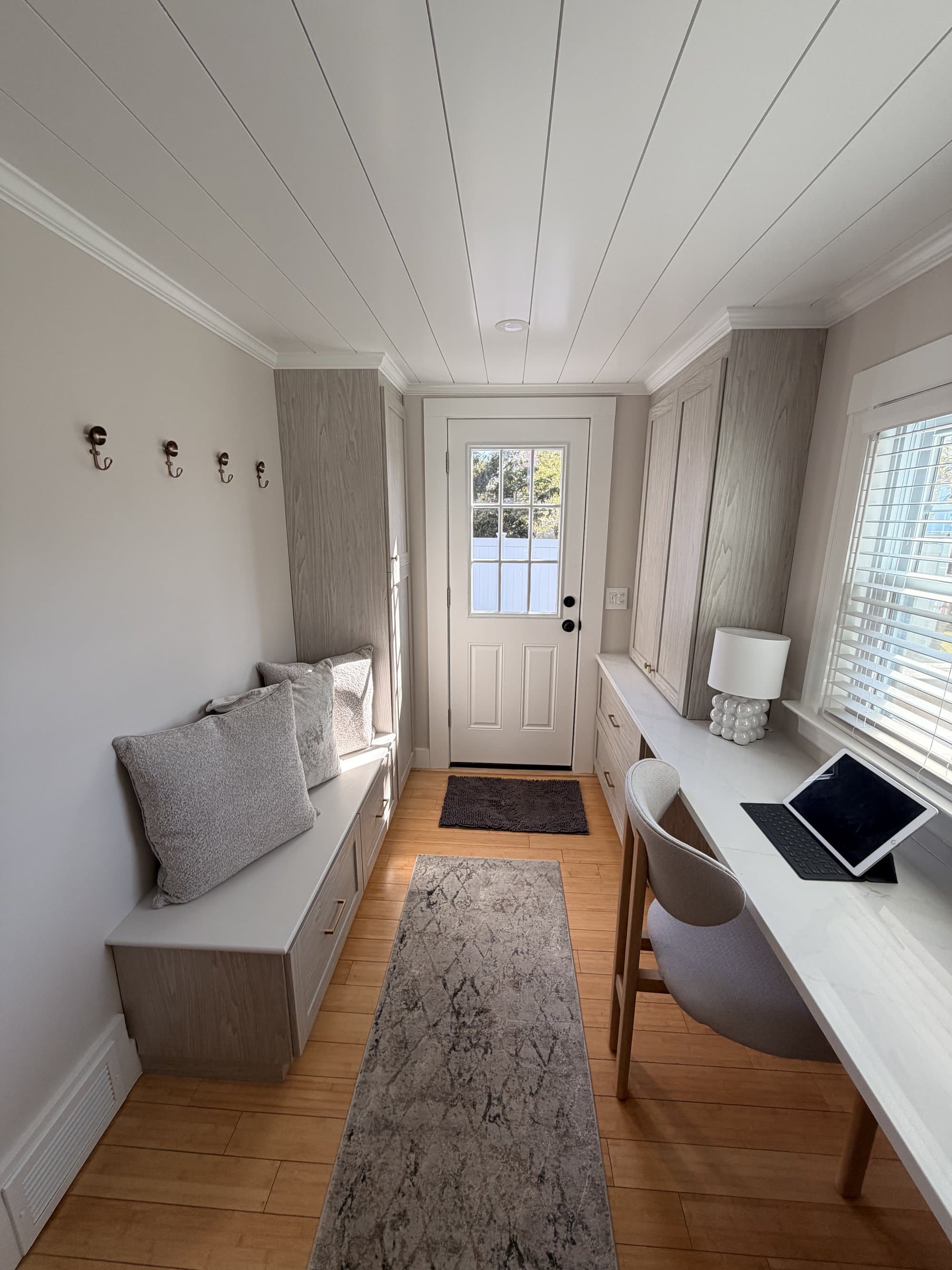 Mirror angle of the finished mudroom showing the cushioned bench, brass coat hooks, new exterior door, and built-in desk run